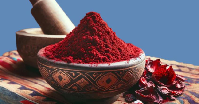Traditional Moroccan ceramic bowl filled with vibrant red Aker Fassi powder, placed next to dried poppy petals and a stone grinder on a patterned cloth background.