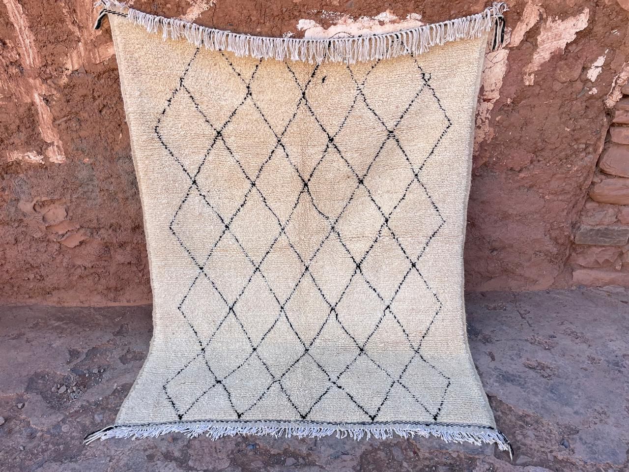 Handmade Moroccan Berber rug in ivory with a black diamond pattern, photographed against a rustic wall, by YAMA Carpet