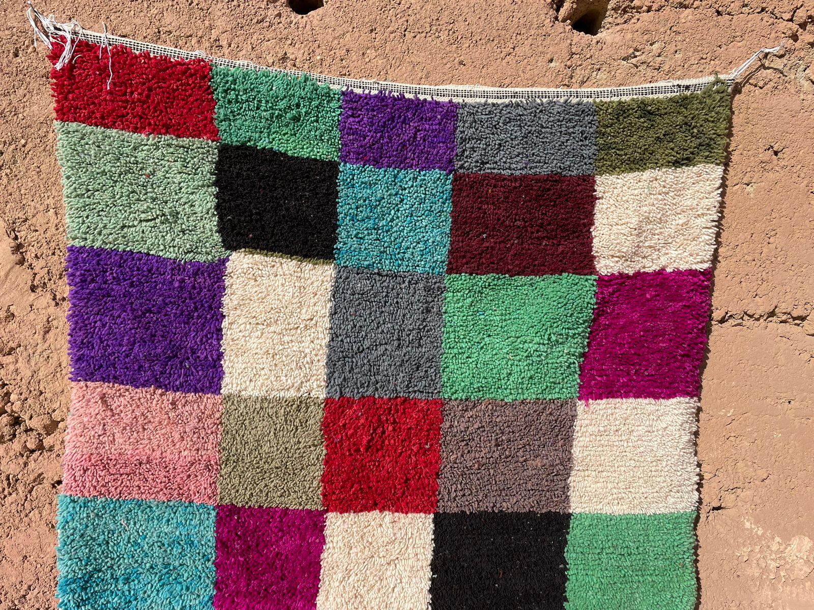 Close-up of handmade Moroccan shag rug with colorful checkered wool pattern displayed outdoors against a mud wall