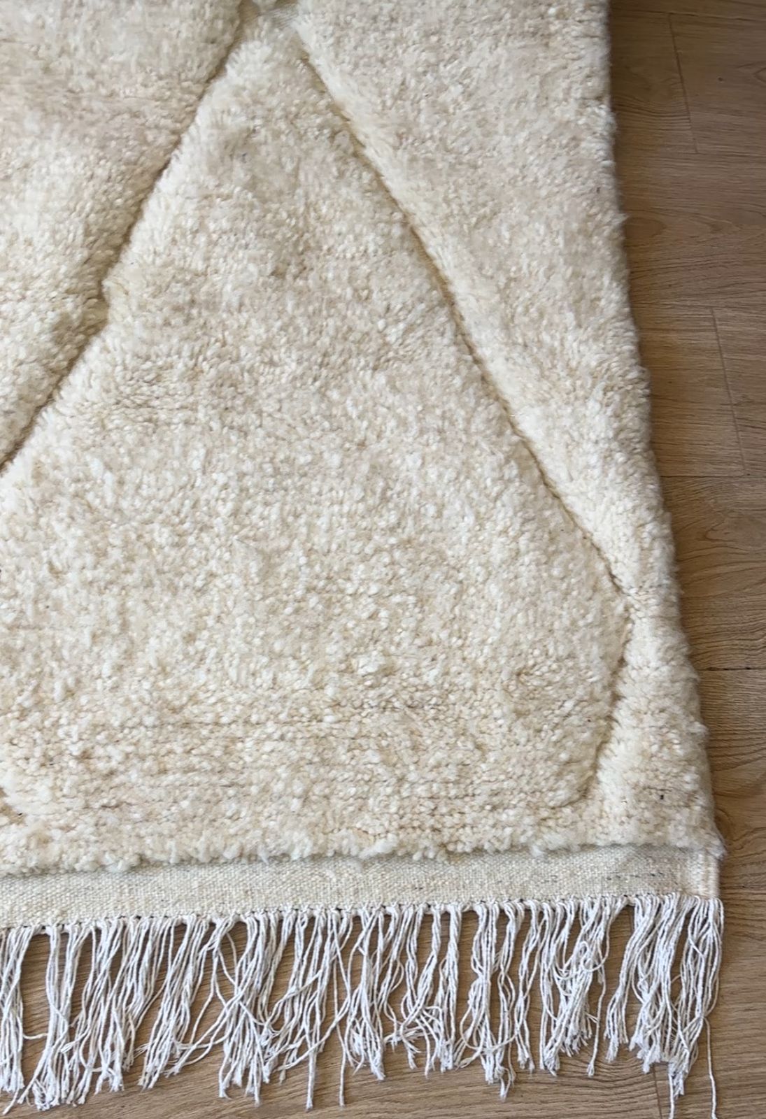 Corner close-up of handmade Moroccan wool rug in cream with stitched diamond design and white fringe on wood floor