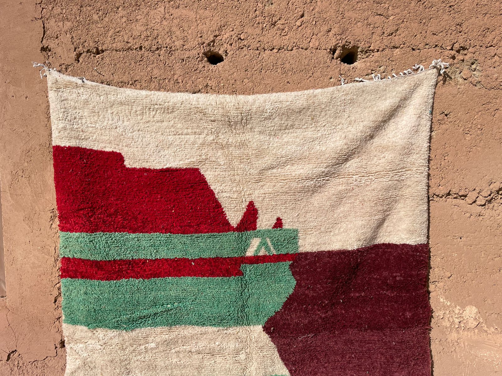 Moroccan handmade rug in red, green, and beige, hanging on earthen wall in natural light
