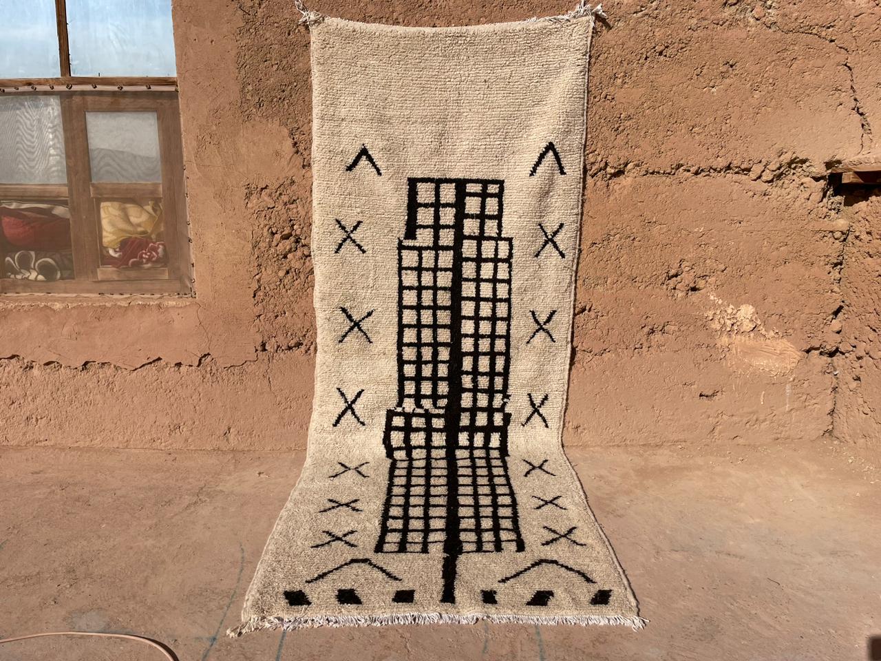 Handmade Moroccan wool rug in beige with bold black grid and tribal details, displayed outdoors against an earthen wall, 5x8 feet